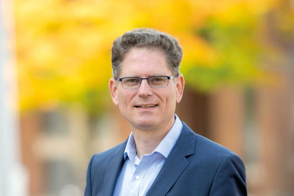 Porträtfoto eines mittelalten grauhaarigen Mannes, in blauem Hemd und Jacket, draußen, im Hintergrund unscharfer gelber Blätterhintergrund